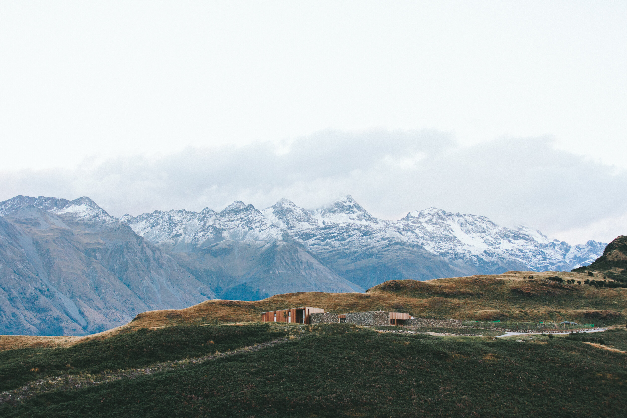 A landscape with snow-capped mountains in the background and rolling green and brown hills in the foreground. There is a modern building on the hill.