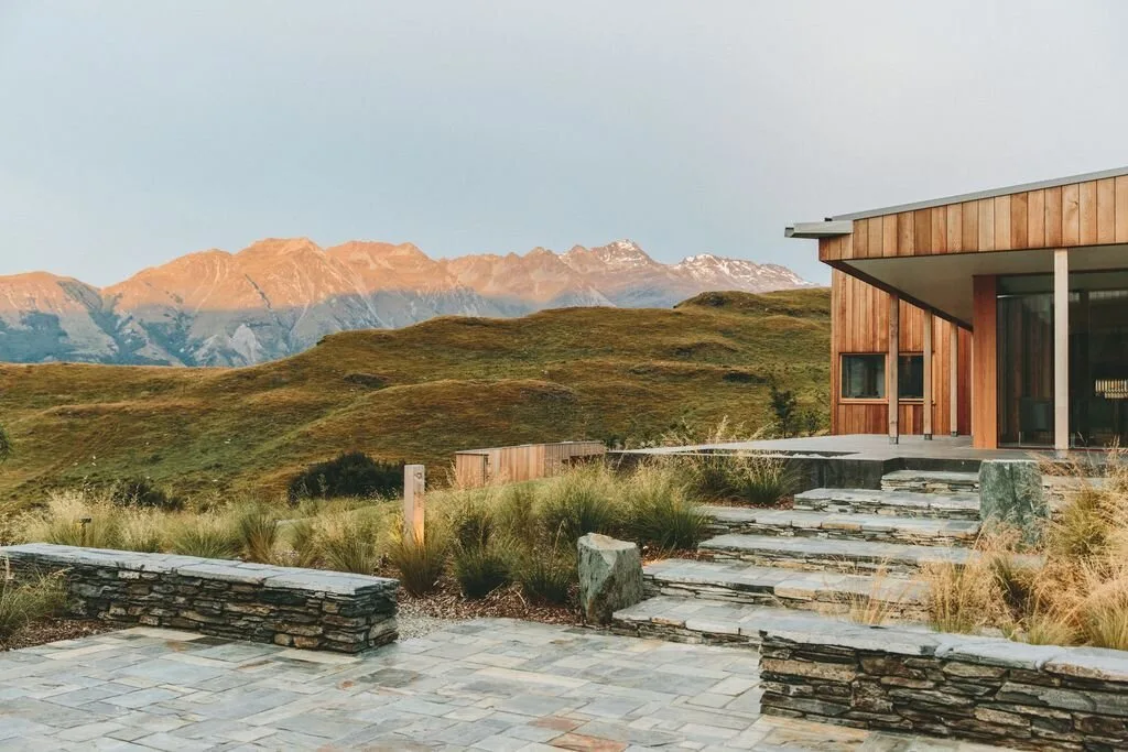 Modern wooden house with stone steps and landscaping, set against green rolling hills and distant mountain range at sunset.