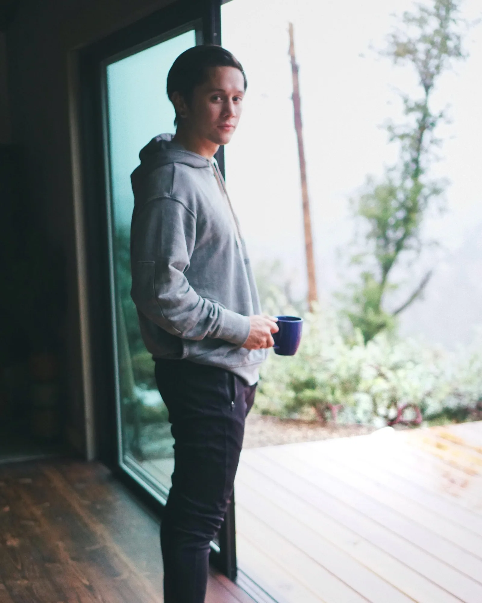 A young man with short dark hair wearing a gray hoodie and black pants stands indoors near a large glass sliding door. He is holding a dark blue mug and looking at the camera. Outside, there is greenery with trees and foggy weather.