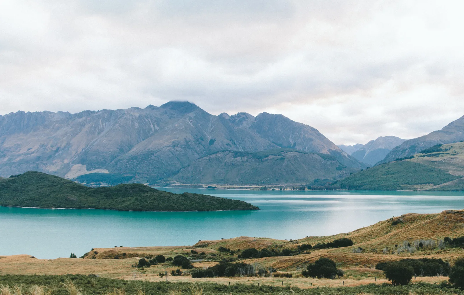 Scenic landscape with a turquoise lake, rolling green hills, and tall mountains in the background under a cloudy sky.