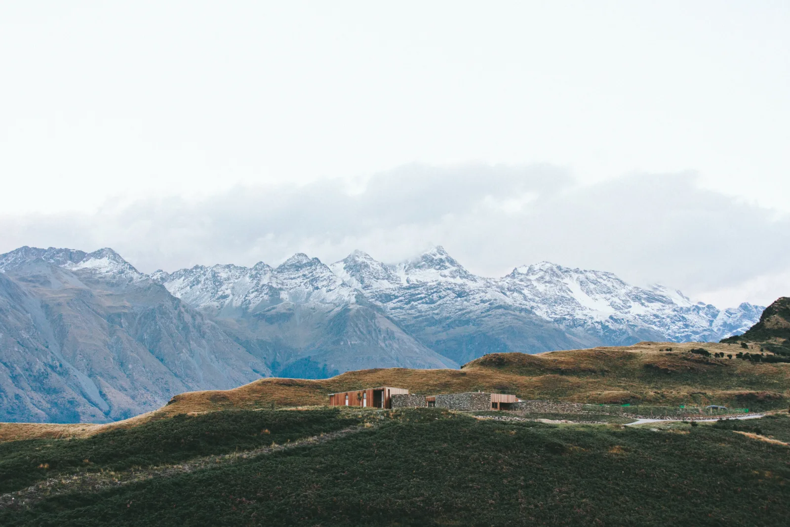 A landscape with snow-capped mountains in the background and rolling green and brown hills in the foreground. There is a modern building on the hill.