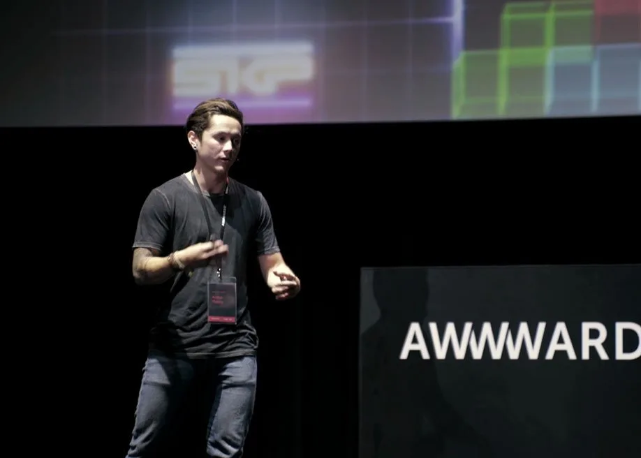 A young man with short dark hair, wearing a black T-shirt and dark jeans, standing on a stage giving a presentation at a conference or event, with a large screen behind him displaying colorful graphics and a sign that reads "AWWWARD."