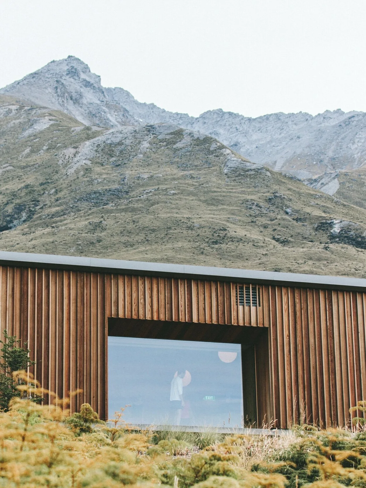 Modern building with wooden exterior and large glass window, mountains in background, and greenery in foreground.